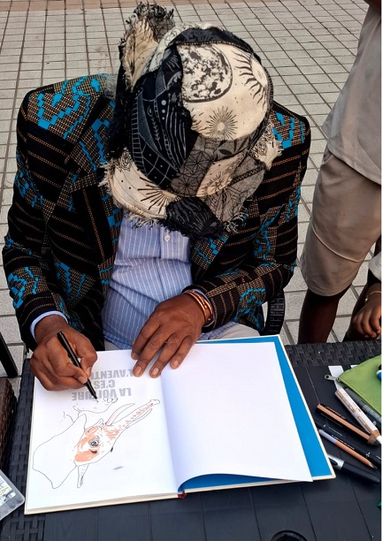 1  : Barly Baruti dédicaçant « La voiture c’est l’aventure »/ Adiac